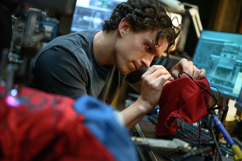 A young man meticulously works on a red garment, focused on sewing or assembling something, with multiple screens showing technical data in the background.